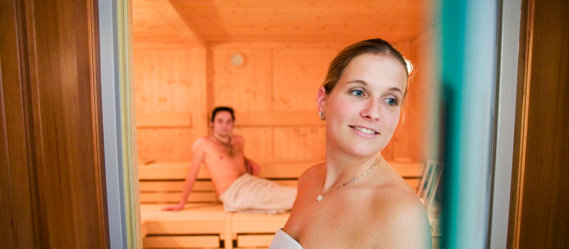 Eine Frau schaut aus einer Sauna durch die Glastür heraus während ein Mann auf einer Saunabank dahinter sitzt