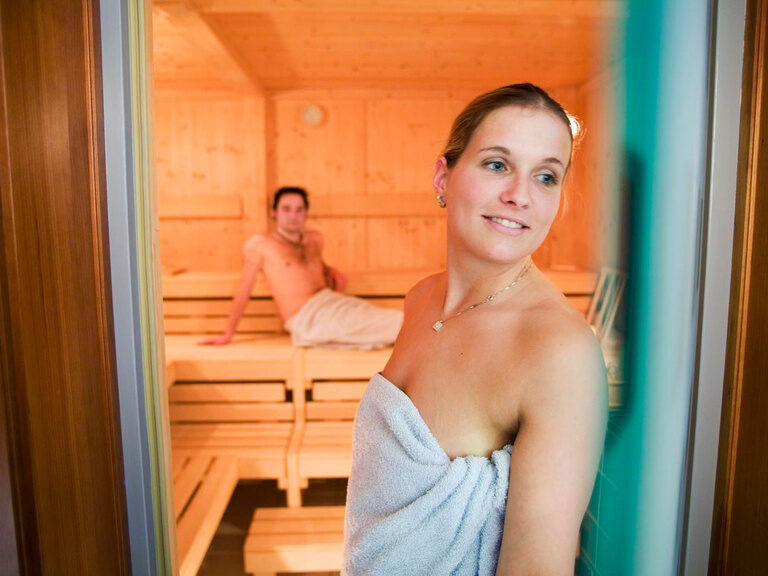 Eine Frau schaut aus einer Sauna durch die Glastür heraus während ein Mann auf einer Saunabank dahinter sitzt